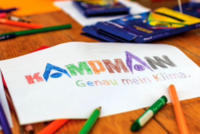 Von Kinderhänden ausgemaltes Kampmann Logo mit Schriftzug Genau mein Klima in vielen verschiedenen Farben, der Tisch liegt voll Stifte